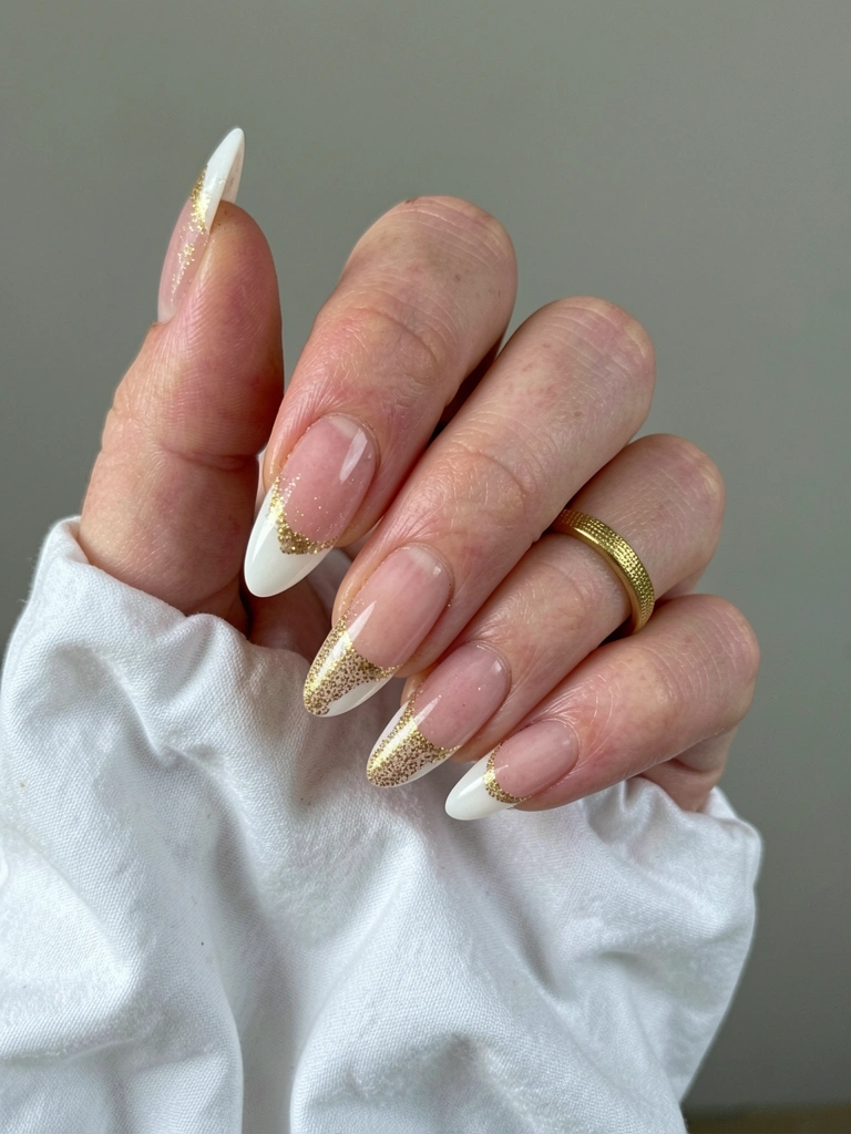 Unhas de gel francesinha com ponta fina e glitter dourado sobre fundo claro.